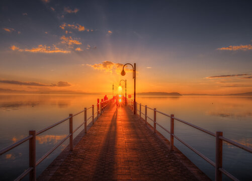 View of a wooden pier extending into the calm waters of Lake Trasimeno at sunset, with warm golden light reflecting on the surface and silhouettes of people walking in Umbria, Italy.
