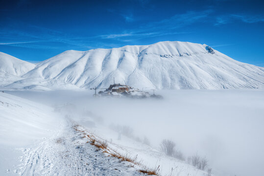 View of the village of Castelluccio di Norcia emerging from a sea of fog with snow-covered mountains in the background in Umbria, Italy.