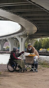 Vertical video of Caucasian senior homeless man talking to Caucasian man with disability in wheelchair near shopping cart. Footage fits social aid, charity, and poverty awareness campaigns