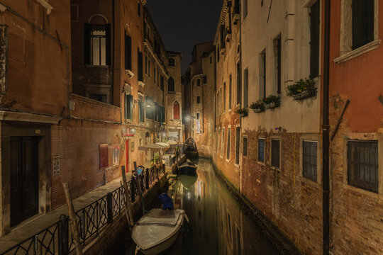 View of a narrow canal at night with historic buildings, reflections in the water, and a small boat moored along the stone embankment in Venice, Veneto, Italy.