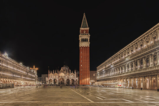 View of St. Mark's Square at night featuring the illuminated St. Mark's Basilica and the Campanile di San Marco surrounded by historic arcades in Venice, Veneto, Italy.