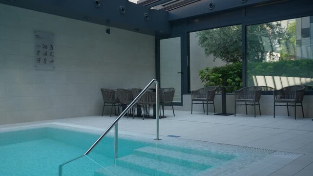 Indoor spa pool features comfortable lounge chairs, dining table and water entry steps. Panoramic glass windows offer a scenic garden view for quiet wellness retreat and healthy relaxation.