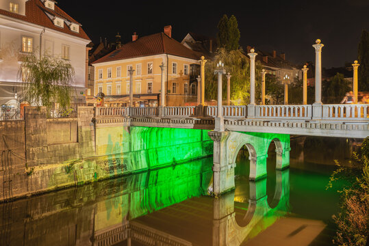 View of the illuminated Triple Bridge crossing the Ljubljanica river at night, featuring historic architecture and reflections in the water in Slovenia.