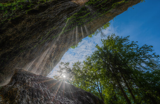 View of a waterfall cascading over a mossy rock cliff with sunlight filtering through the surrounding green trees against a blue sky in Slovenia.