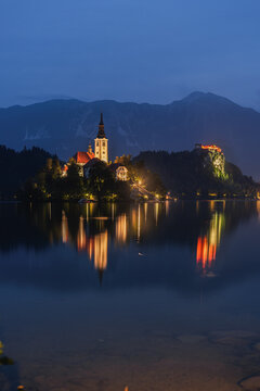 View of the illuminated Bled Island with its church and the distant Bled Castle reflecting on the calm waters of Lake Bled, Slovenia.