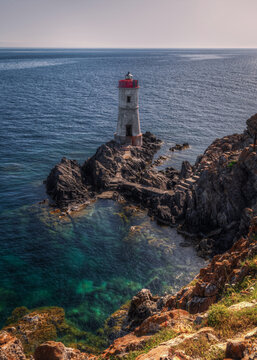 View of a lighthouse with a red lantern room perched on rugged coastal rocks surrounded by clear turquoise sea in Sardinia, Italy.