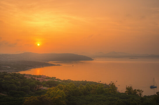 View of the sun setting over the coastal landscape with lush greenery, calm sea waters, and a lone sailboat in the distance, Sardinia, Italy.