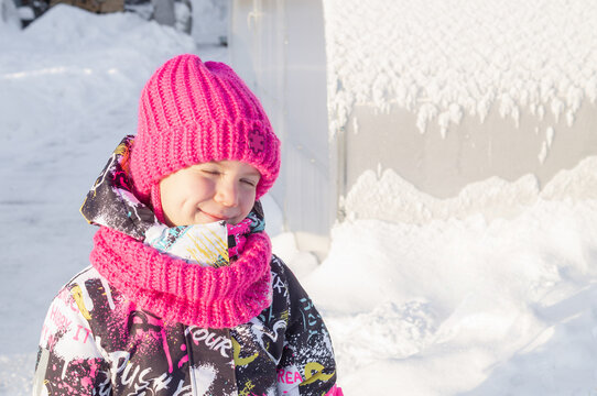 Cute little girl smiling and squinting in bright winter sun outdoors. Happy child in vivid pink knitted hat and colorful warm outerwear. Childhood, winter lifestyle and kids fashion. Copy space.