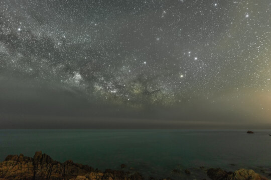 View of the Milky Way galaxy stretching over the rocky coastline and dark sea in Sardinia, Italy.