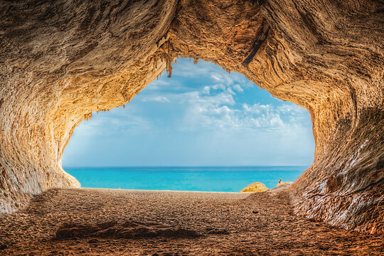 View of a natural sea cave with rocky walls and a sandy floor looking out over the turquoise Mediterranean sea in Sardinia, Italy.