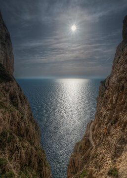 View of dramatic rocky cliffs framing the sunlit sea with a narrow stone path carved into the rock face in Sardinia, Italy.