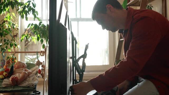 Young father carefully installing a new flat screen television on a wall mount bracket while his infant baby is resting in a bouncer in a sunlit living room