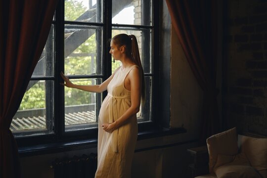 A pregnant woman stands by a window in her home, admiring the view outside. She appears reflective and serene, dressed in a simple, flowing dress