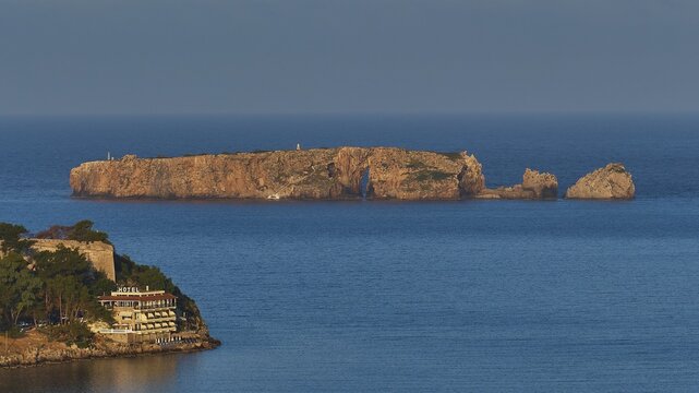 Small island near coast with hotel and trees against blue sky, morning light, Tsichli Baba island, Navarino Bay, Pylos, Peloponnese, Greece