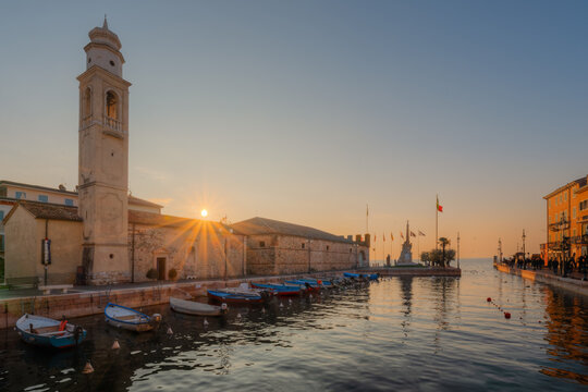 View of the historic Church of San Nicolo with its bell tower overlooking the small harbor filled with wooden boats during a golden sunset in Lazise, Veneto, Italy.