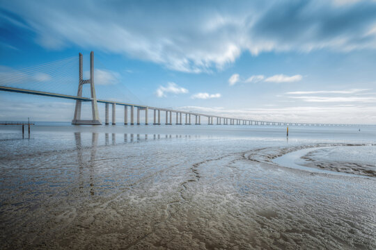 View of the Vasco da Gama Bridge spanning the Tagus River with mudflats in the foreground under a blue sky in Lisbon, Lisbon, Portugal.