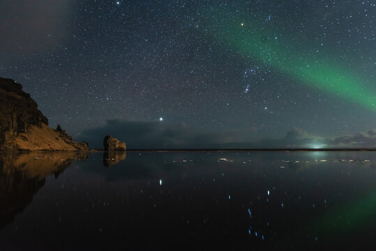 View of the aurora borealis illuminating the night sky above a calm, reflective body of water with dark rocky cliffs in Iceland.