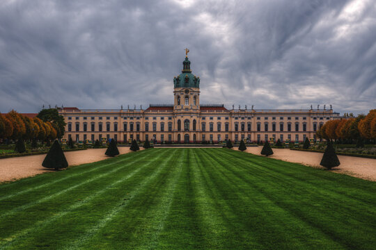 View of Charlottenburg Palace with its manicured green lawn, symmetrical topiary trees, and historic baroque architecture under a dramatic cloudy sky in Berlin, Berlin, Germany.