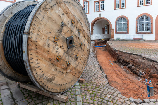 Large wooden cable reel stands beside excavated trench during utility installation in historic courtyard. Underground infrastructure work shows conduit, cobblestones, soil and renovation site