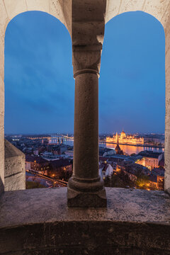 View of the illuminated Hungarian Parliament Building across the Danube River, framed by the stone arches and columns of Fisherman's Bastion in Budapest, Hungary.