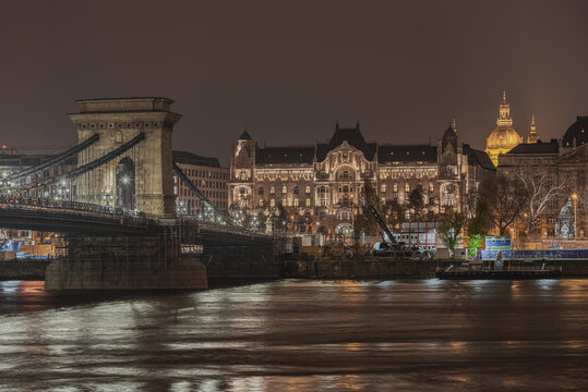 View of the illuminated Szechenyi Chain Bridge spanning the Danube River at night with historic architecture in the background in Budapest, Hungary.