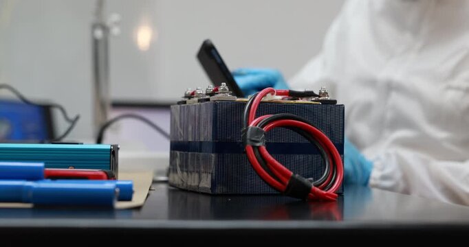 Technician in protective clothing works with a compact battery pack and red cables on a laboratory table, electronics repair and energy storage testing.
