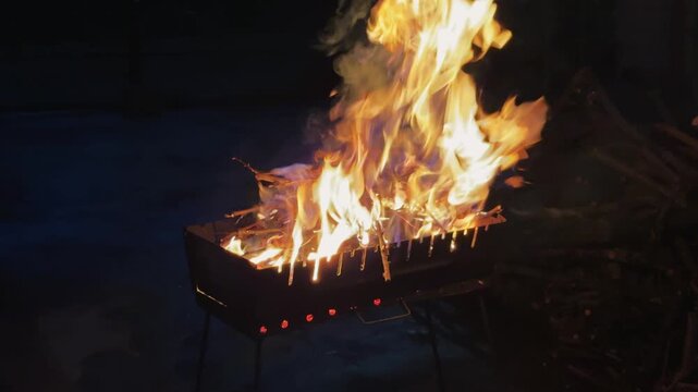 Burning branches with bright flames in a metal brazier outdoors at night with smoke rising from the fire