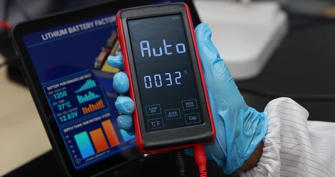 Gloved technician holds a digital battery tester auto mode and temperature beside a tablet with lithium battery health charts.
