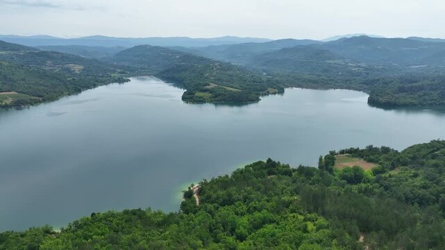 Turquoise water of Butoniga reservoir reflects white clouds in Istria region. Drone flies over artificial lake surrounded by dense green forests and rolling hills near Motovun.