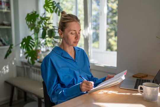 Concentrated positive woman freelancer planning day and tasks while sitting at table in kitchen at home, confident female businesswoman taking notes in notebook, morning remote work daily routine.