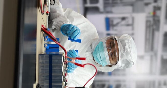 Technician in clean room protective suit, mask and gloves checks electrical equipment with red test leads in a modern industrial laboratory.