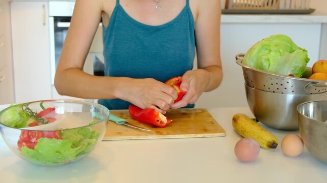 Unrecognizable woman slices sweet red bell pepper into small pieces on wooden board. Glass bowl with mixed green lettuce and red tomatoes stands on white table during healthy cooking.