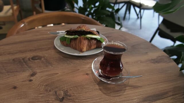 Glass of dark Turkish tea stands on wooden table next to delicious croissant sandwich. Cozy cafe interior with green plants creates relaxing atmosphere for breakfast or lunch.