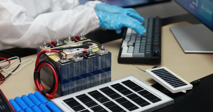 Technician in protective gloves works at a computer beside battery cells, cables and small solar panels during renewable energy research.