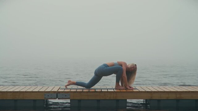 Athletic blonde woman in sportswear performs the elbow to instep, world's greatest stretch on a wooden deck beside a misty lake, promoting mobility, flexibility and active outdoor wellness.