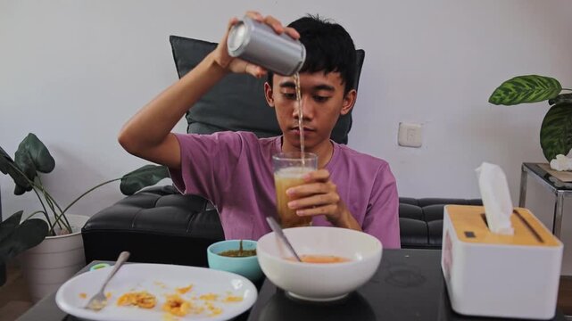 Young Asian man pouring canned tea into a glass while eating batagor at home