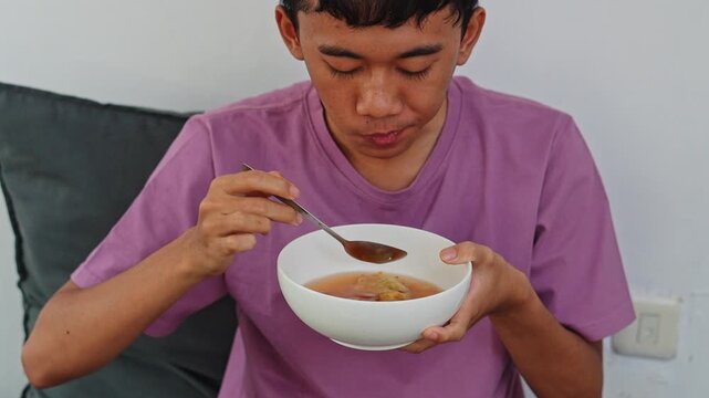 Young man holding white bowl and sipping soup while eating batagor at home