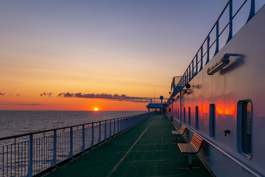 Empty cruise ship deck during a beautiful golden sunrise over the calm sea. Features wooden benches, metal railings, and vibrant sun reflection on the boat facade at dawn, captured at Baltic sea.