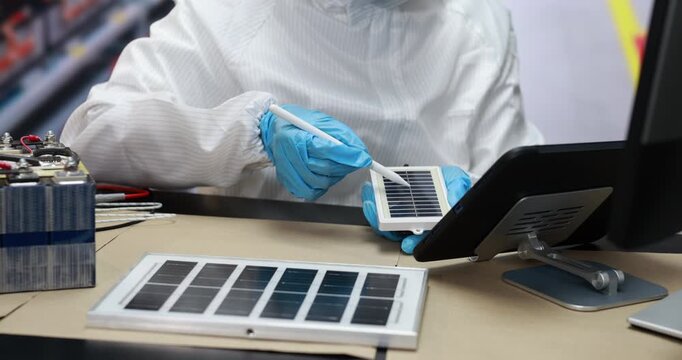Technician in protective clothing and blue gloves tests a small solar panel at a workbench with sample cells, tools, and computer equipment.