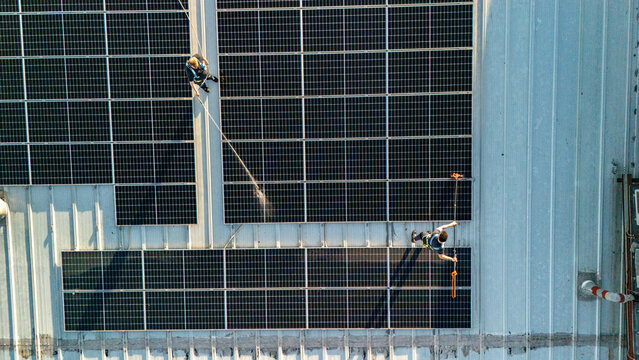 Workers are on the rooftop of a large building, installing solar panels. They are using tools and safety gear while carrying out their tasks.