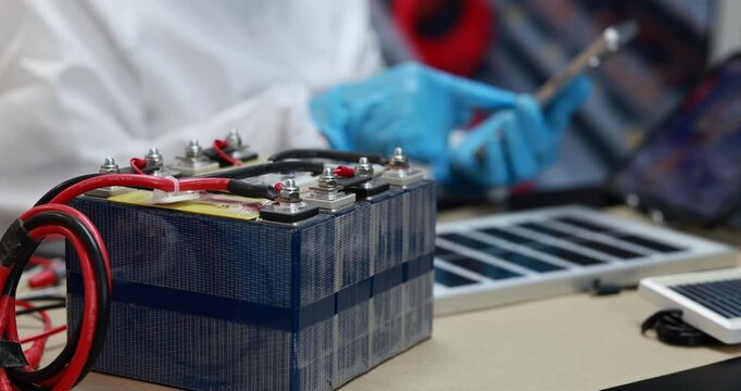 Close-up of a battery pack with red and black cables on a workbench, while a technician in blue gloves uses tools in a workshop.