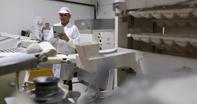 Female food technician in a white uniform supervising a commercial dough making machine on a conveyor belt, holding a tablet to check the automated production line process in a factory
