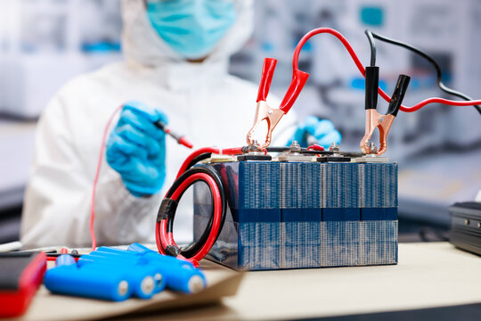 Scientist in protective gear tests a large battery pack with electrical clips and wires in a modern laboratory, with blue batteries in the foreground.