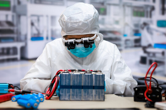 Technician in protective clean room clothing assembles a rechargeable battery pack at a modern laboratory workbench, with wires, tools, and equipment in the background.