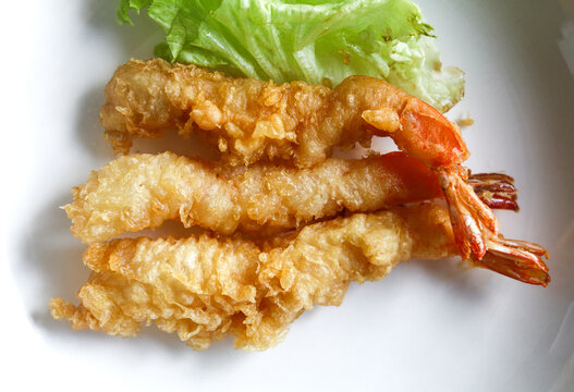 Closeup view of crispy battered ebi tempura pieces with red tail ends arranged neatly on a white dish with a lettuce garnish.
