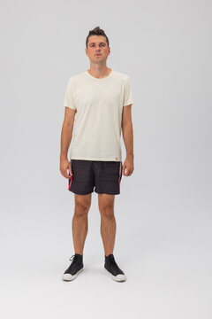 A young Caucasian man in a casual full-body pose, wearing a minimalist cream t-shirt, athletic black shorts with red piping, and high-top black sneakers on a studio background.