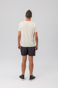 Full length rear view of a young man wearing a plain cream t-shirt, dark shorts, and black high-top sneakers. Standing against a neutral studio background. Perfect for apparel design mockup.