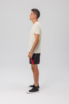 Full-length side view of a young man standing in profile, wearing a cream t-shirt, black sports shorts with red trim, and sneakers against a grey background. Clean studio mockup photo.