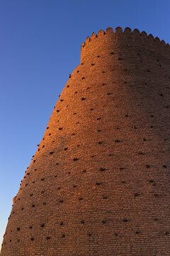 Massive brick tower of the Ark Fortress in Bukhara bathed in golden sunset light against a clear blue sky. Ideal for travel guides, history publications, and Central Asian cultural content.