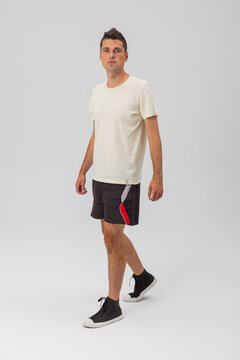 Full-length three-quarter view of a young man walking in a studio setting, dressed in a plain cream t-shirt, black sports shorts, and high-top sneakers against a light grey background.